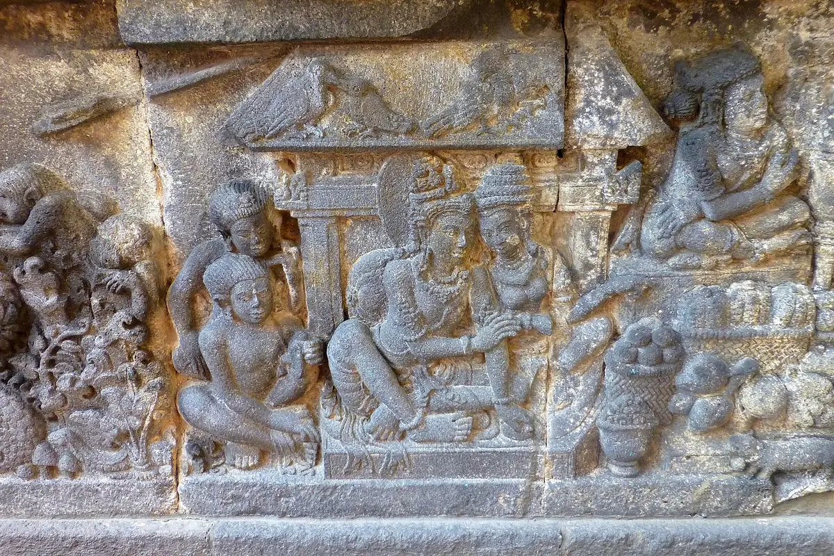 A carved stone relief panel from the Shiva temple at Prambanan showing seated figures under a small roof, with monkeys at left and baskets of fruit at right