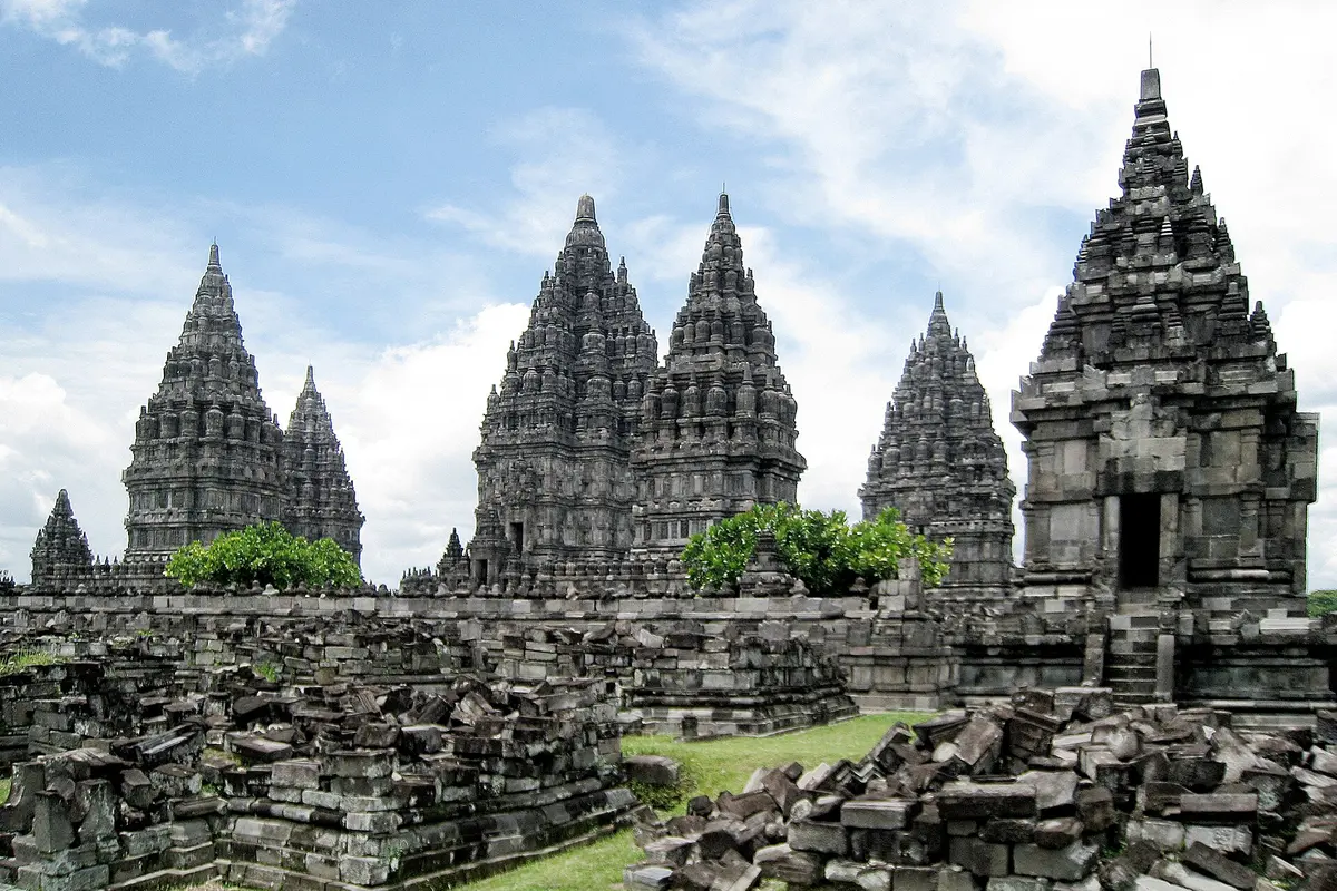 Prambanan's tall pointed temples rising behind rows of fallen grey stone blocks and a low wall