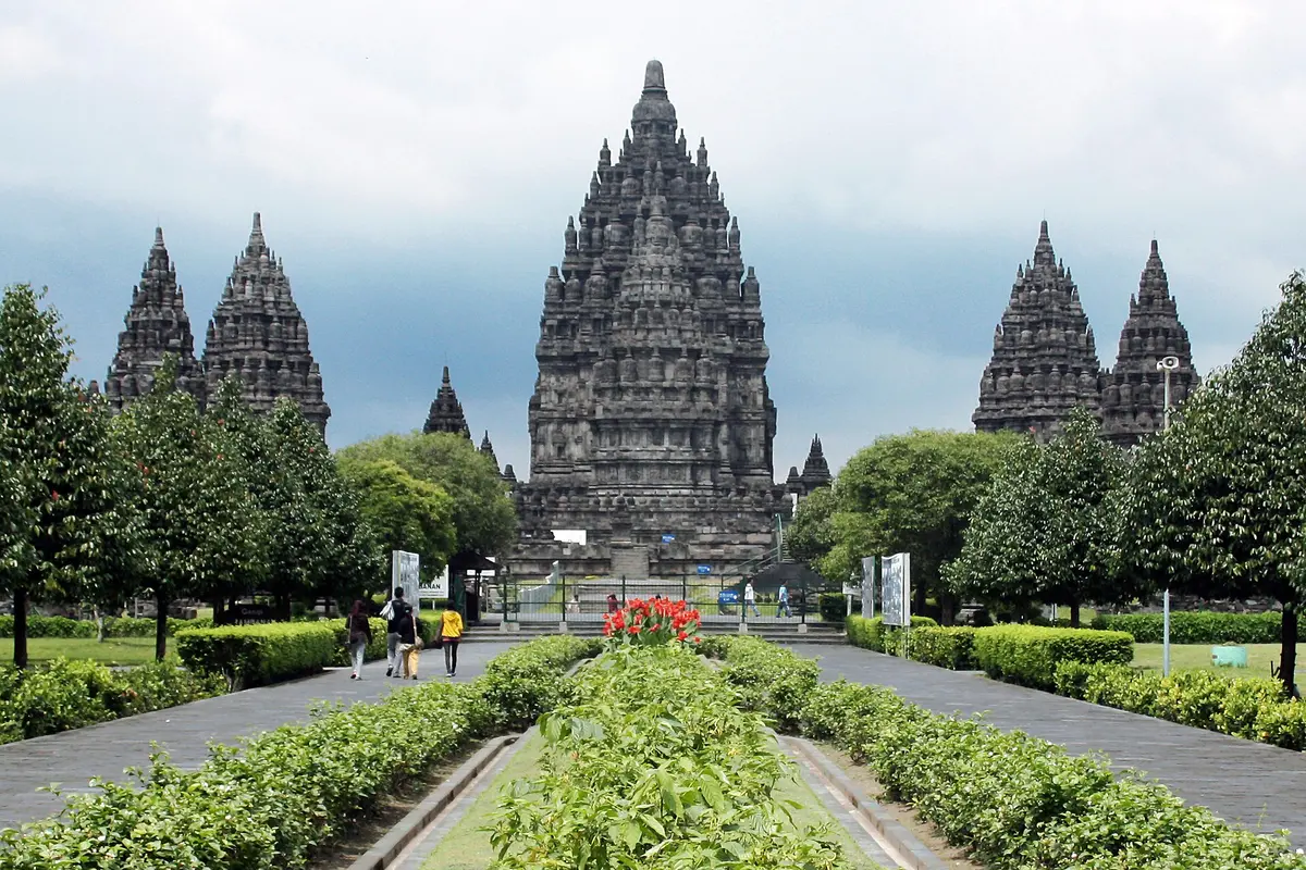 The central Shiva tower of Prambanan with two towers on each side, seen along a paved path lined with hedges and trees
